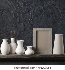 White Ceramic Vases And Wood Photoframe On Distressed Wooden Shelf Against Rough Plaster Black Wall. Home Decor.