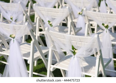 White Chair With A Small Flower. White Decorated Chairs On A Green Lawn. Chairs Set In Rows For The Wedding Ceremony. They Are Decorated For The Festive Event. Chairs Are On The Green Lawn Outside.