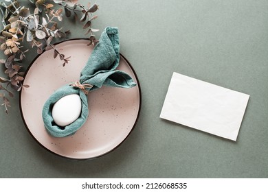 A white chicken egg is wrapped in a napkin in the form of a rabbit on a plate, a piece of paper for writing and a branch of eucalyptus on a green background. Eco friendly happy easter. Top view.
