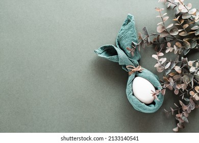 A white chicken egg is wrapped in a napkin in the form of a rabbit and a branch of eucalyptus on a green background. Eco friendly happy easter. Top view.