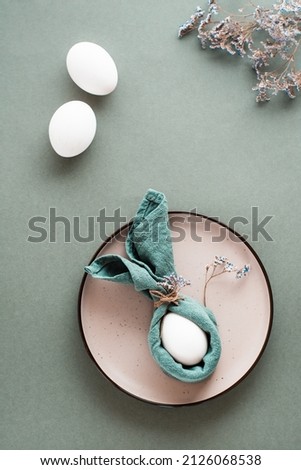 A white chicken egg is wrapped in a napkin in the form of a rabbit on a plate and gypsophila on a green background. Eco friendly happy easter. Top and vertical  view.