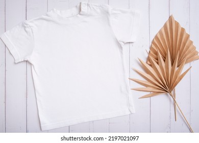 White Children's T-shirt Mockup For Logo, Text Or Design On Wooden Background With Palm Leaves Top View.