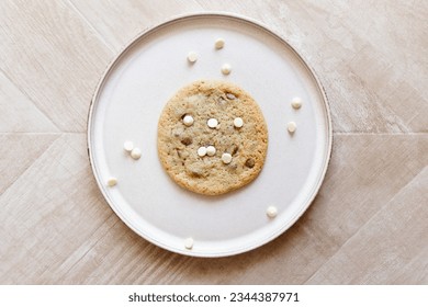 White chocolate and milk chocolate chip cookie on a cream ceramic plate with chocolate chips scattered around. The background is a chevron wood texture. - Powered by Shutterstock