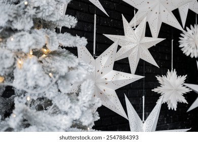 A white Christmas tree adorned with lights stands beside large white star decorations hanging against a dark wall, creating a cozy winter atmosphere perfect for the holiday season. - Powered by Shutterstock