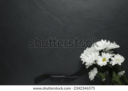 White chrysanthemum flowers and ribbon on black table, top view with space for text. Funeral symbols