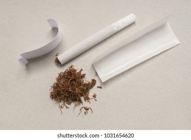 White cigarette paper, cartonboard filter tip, portion of tobacco, and hand rolled cigarette on grey cardboard background.