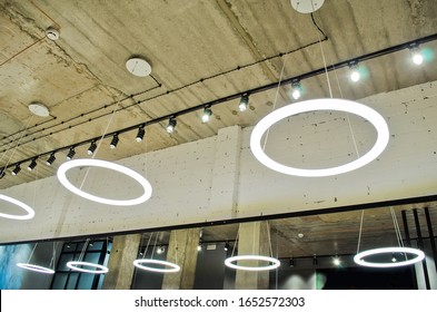 White Circular Ring Shaped Lamps Hang Under A Concrete Ceiling With External Wiring And Black Fixtures, With Mirror Reflection