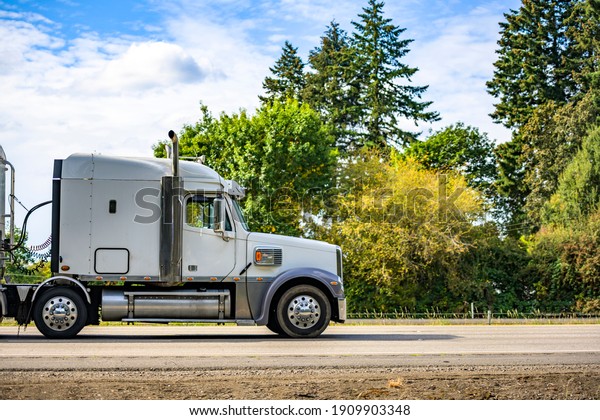White classic big rig industrial professional semi\
truck with vertical pipes and horns on the roof transporting cargo\
in covered bulk semi trailer running on the flat highway road with\
green trees