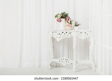 A white classic table with flower vase in front of white curtain and white window