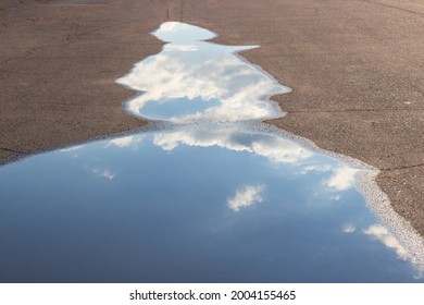 White clouds and sky are reflected in a puddle on the asphalt on the road.