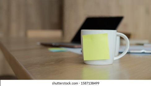 A white coffee cup with a blank yellow Sticky Note on the cup. Resting on the desk and laptop is placed on the back. - Powered by Shutterstock