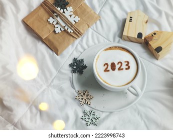 White coffee cup with number 2023 over frothy surface flat lay on the bed with white blanket, wooden house model, paper gift bag, snowflake sign. Home 2023, Happy new year theme (top view, copy space) - Shutterstock ID 2223998743