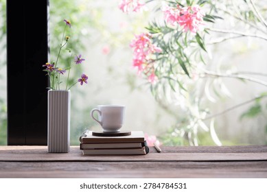 White coffee cup on stack of old notebook with pink flowers in a white vase, in front of window