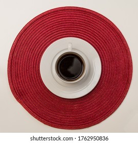 White Coffee Cup With White Saucer Standing On The Red Round Straw Table Mat