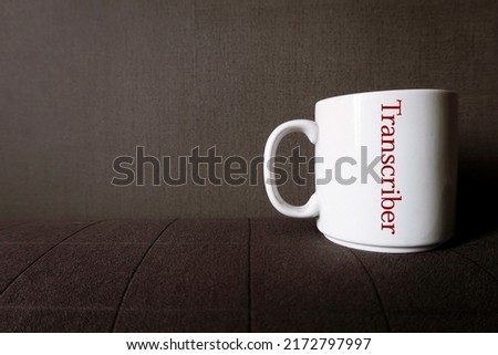 White coffee mug cup on copy space dark sofa with text inscription TRANSCRIBER, means Transcriptionist or a person who listen to recorded audio video or other media and transcribe to writing words