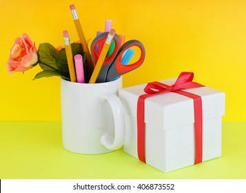 White coffee mug filled with markers, pencils, scissors and a silk rose with a gift box on a green and yellow background good for secretary's day, or administrative professionals day