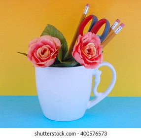 A white coffee mug filled with office supplies like pencils and a pair of scissors with a couple of silk roses on a blue and yellow background. Good for administrative professional'??s day in April