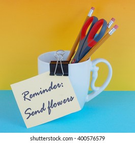 A white coffee mug filled with pencils and a pair of scissors with a binder clip holding reminder on a blue and yellow background. Good for administrative professional's day in April