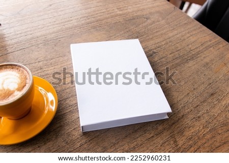 A white colored hardcover book with a wooden textured table as the background and a coffee near it, hardcover mockup template image