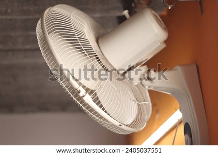 A white colored Wall revolving fan or table fan on an orange wall with tube light in background