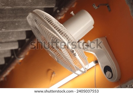 A white colored Wall revolving fan or table fan on an orange wall with tube light in background, selective focus