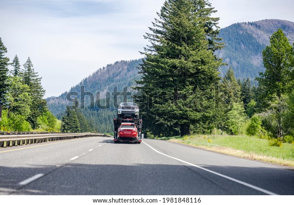 White compact big rig industrial car hauler semi\
truck transporting cars on the hydraulic modular two level semi\
trailer running on curving highway road with green mountain forest\
in Columbia Gorge