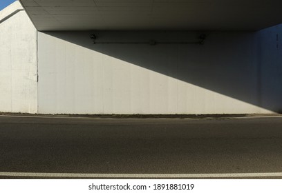 White concrete wall under an underpass divided in two obliquely by a shadow. Asphalt road in front. Background for copy space.