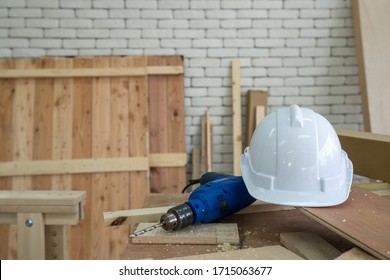 The White Construction Helmet And The Blue Electric Drill Were Left Aside During Lunch Time Brake. A Desk Full Of Hand Tools And Wood Piles. Morning Work Atmosphere In The Workshop Room.