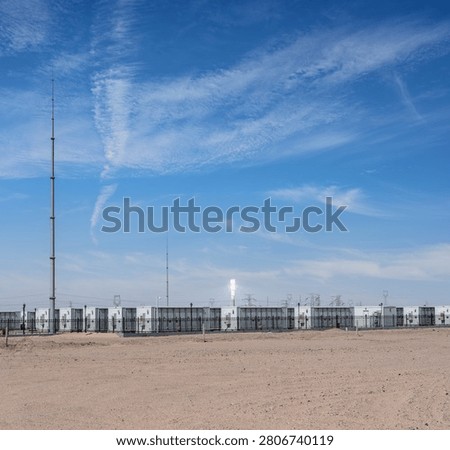 White containerized battery storage units line a fenced site in the arid Gobi desert; a bright solar thermal tower glows in the distance under a vivid blue sky.  