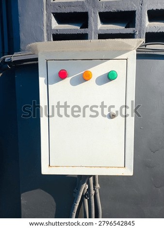 White control box mounted on dark wall. Red, yellow, and green buttons stand out clearly. Sunlight casts soft shadows across the surface. Metal latch and pipe connections show industrial use