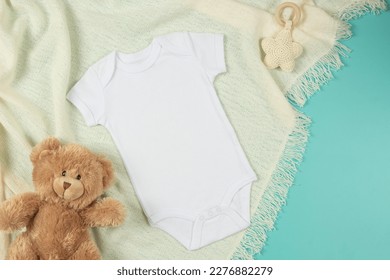 White cotton baby short sleeve bodysuit, teddy bear and natural wooden toy on beige blanket throw background. Infant onesie mockup. Blank gender neutral newborn bodysuit mock up template. Top view
