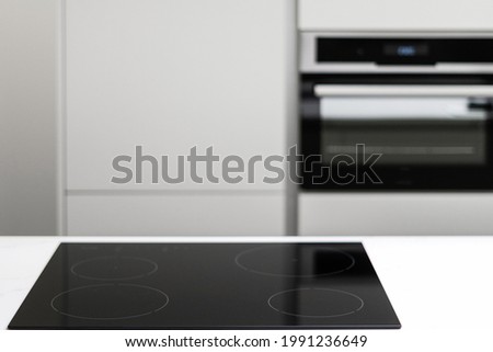 White countertop with glossy built in ceramic black induction stove, big white cupboard with built-in electrical oven in empty kitchen with nobody in sight, blurred background