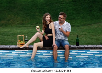 A white couple in love hugs on the edge of the pool. Feet in the water. They smile and laugh. They drink wine from a glass. Eats an apple. Green grass background. Rest in the garden. Bottle and glass. - Powered by Shutterstock