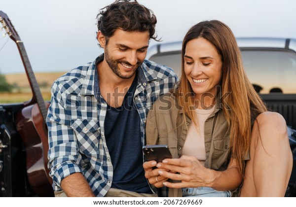 White couple smiling and using cellphones while\
sitting in car trunk\
outdoors