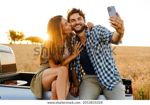 White couple taking selfie photo on cellphone
while sitting on car trunk
outdoors