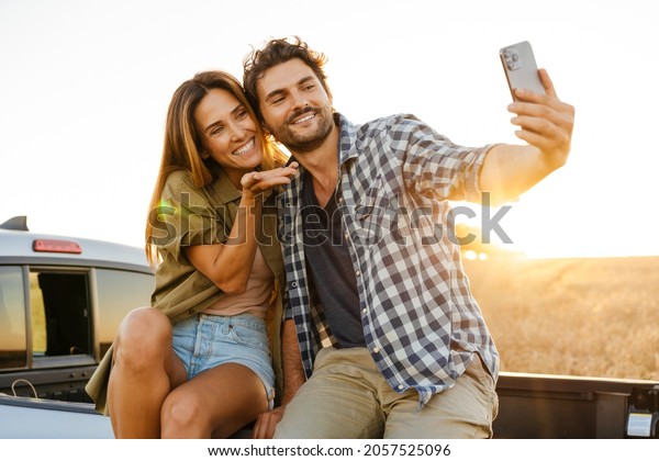 White couple taking selfie photo on cellphone\
while sitting on car trunk\
outdoors