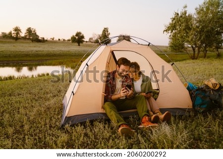 White couple using cellphones and sitting in tent during camping together on summer day