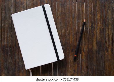 White cover notebook with black pencil on rustic wooden table flat lay photo. Closed notebook with blank cover flat lay photo. Notepad on table top view. Sketchbook banner template. Art logo mockup