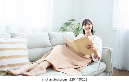 White Cozy Bed And Asian  Beautiful Girl, Reading A Book, Concepts Of Home And Comfort, Place For Text.Book Worm Love Read Book .Charming Woman On Cozy Sofa Interior.Vacation Activity In Home.