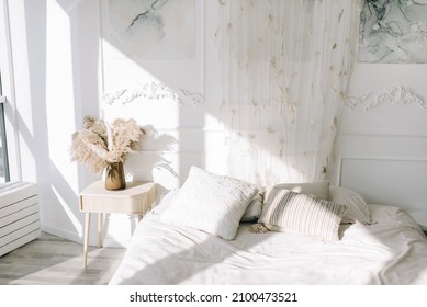 White cozy bedroom  where the sun shines from the window. White bedding, dried flowers in a vase on a bedside table for decoration.