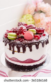 White Cream Mousse Cake Decorated With Pomegranate, Colorful Macaroons And Chocolate Glaze On White Background With Defocused Flowers