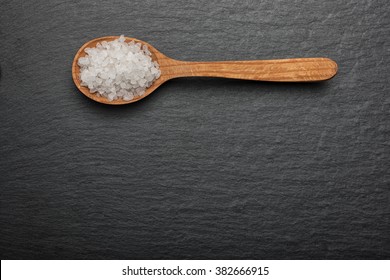  White Crystals Sea Salt On Wooden Spoon On Black Stone Background, Top View