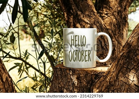 white cup of coffee with hello October text on Tree trunk wood Podium through olives tree