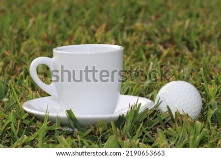 White cup of coffee next to a golf ball, on a green lawn