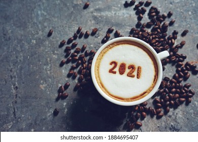 White Cup With Coffee And Number 2021 Over Frothy Surface Flat Lay On Grey Cement Background With Coffee Beans And Space For Text. New Year New You, Holidays Food Art Theme Happy New Year 2021.