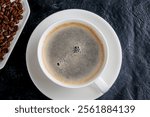 A white cup of freshly brewed coffee sits on a saucer, accompanied by scattered coffee beans on a dark surface, creating an inviting atmosphere.