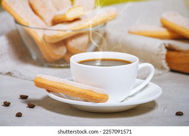 A white cup of hot aromatic coffee with steam and traditional Traditional italian savoiardi biscuits or ladyfingers cookie on a saucer and in a glass bowl. Selective focus