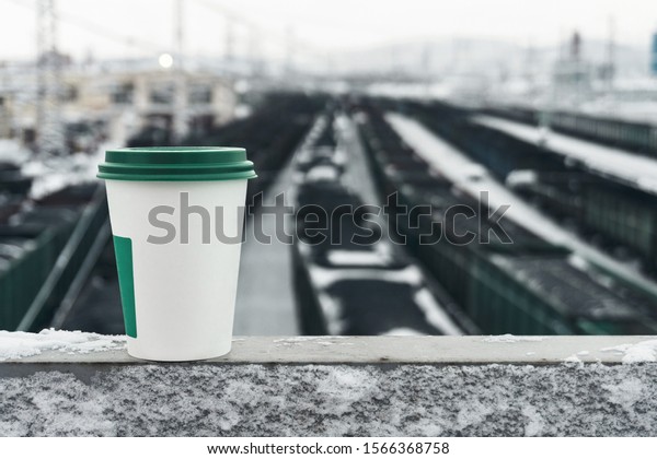 a white cup for hot coffee with a green\
lid stands on the railing of the bridge over the railway station\
with dark freight cars on a frosty winter\
day
