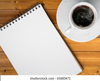 White Cup Of Hot Coffee And White Sketch Book On Wood Table