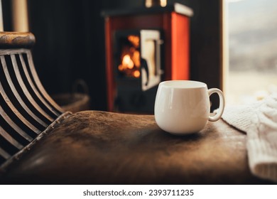 White cup with hot tea with burning fireplace on the background in cozy log cabin.